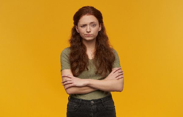 Unhappy looking girl, distrustful redhead woman with long hair. Wearing green t-shirt. People and emotion concept. Holding arms crossed. Watching at the camera, isolated over orange background